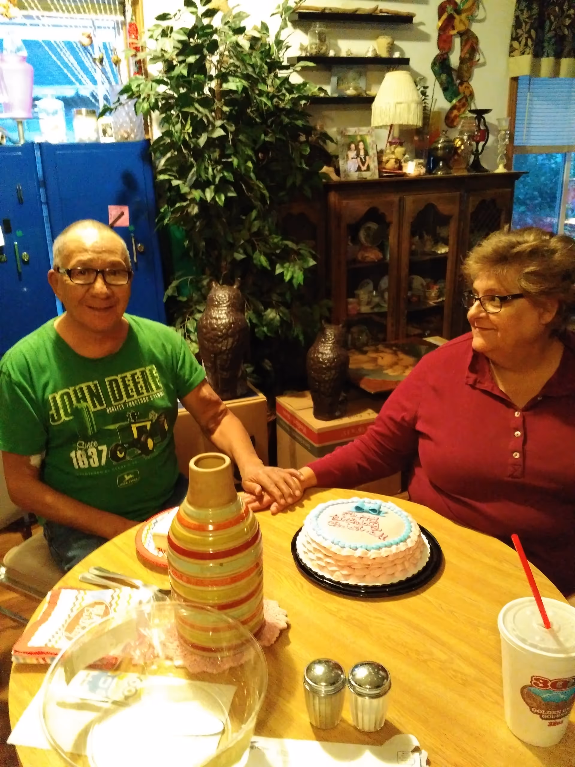 Two people sitting at a wooden dining table holding hands. On the table, there is a small decorated cake, a striped vase, salt and pepper shakers, a large drink cup with a red straw, and some napkins. Behind them, there is a cabinet with decorative items and a large green plant.