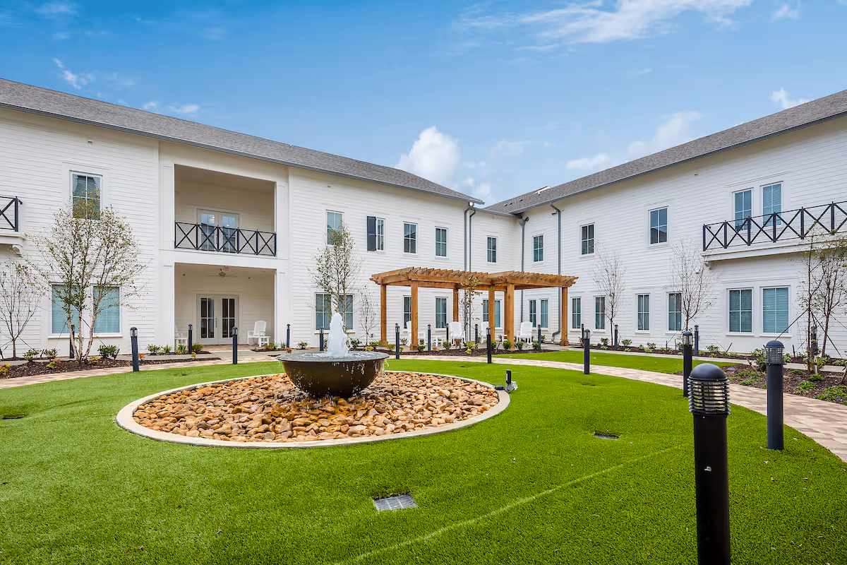 Courtyard of a two-story white senior living building with a central fountain, pergola, lawns and walkways.