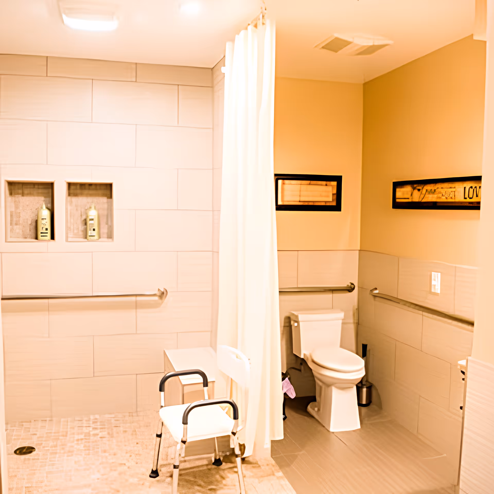 Accessible bathroom with a walk-in shower featuring two built-in shelves with shampoo bottles, a white shower curtain, a white shower chair with black handles, a toilet with grab bars on the walls, and two framed pictures on the wall.