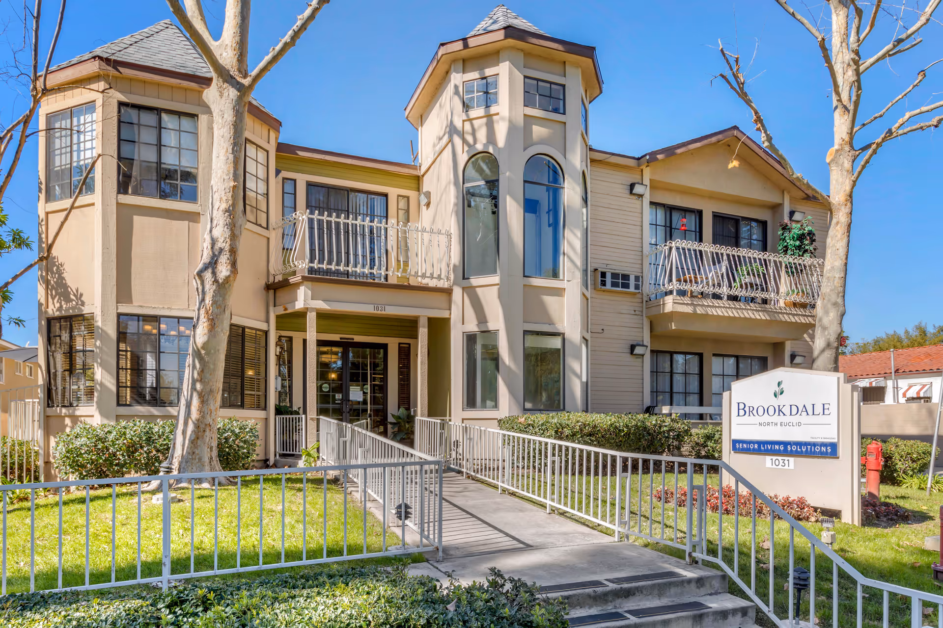 Exterior front view of Brookdale North Euclid senior living facility showing a two-story building with large windows, balconies, and a ramp leading to the entrance. The building is surrounded by green grass, bushes, and leafless trees under a clear blue sky.