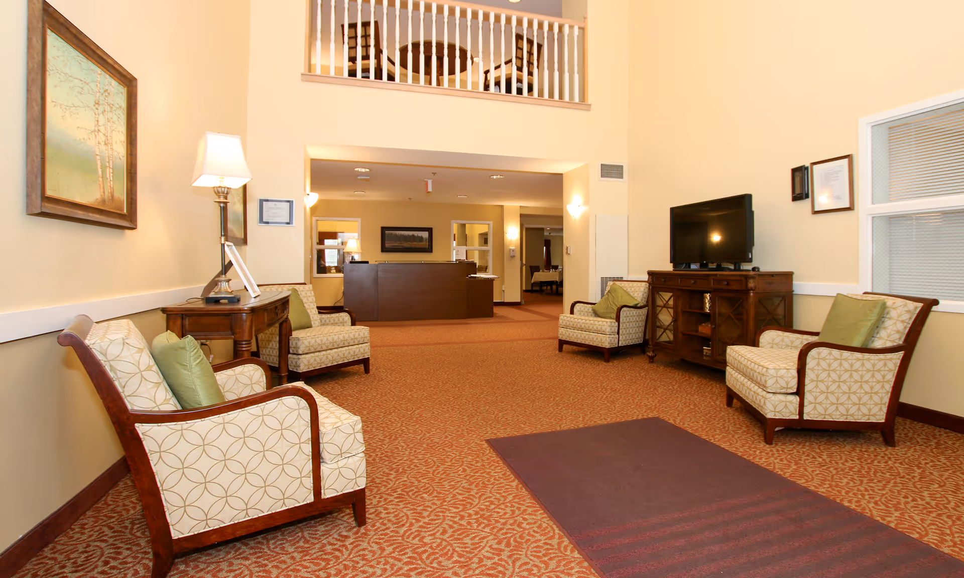 Spacious senior living lounge with upholstered chairs, a TV console, and a reception desk visible in the background.