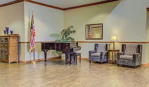 A spacious room with a polished wooden floor featuring a black grand piano with a matching bench, two upholstered armchairs with striped cushions, a small side table with a lamp, an American flag on a stand, a wooden cabinet with glass doors, a potted plant, and a wall mirror reflecting part of the room.