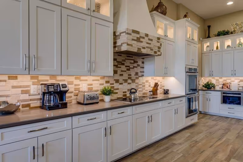 A modern kitchen with white cabinets, a tiled backsplash in neutral tones, a coffee maker, toaster, kettle, and various decorative items on the countertops and shelves. The floor is wooden, and the kitchen has built-in ovens and ample storage space.