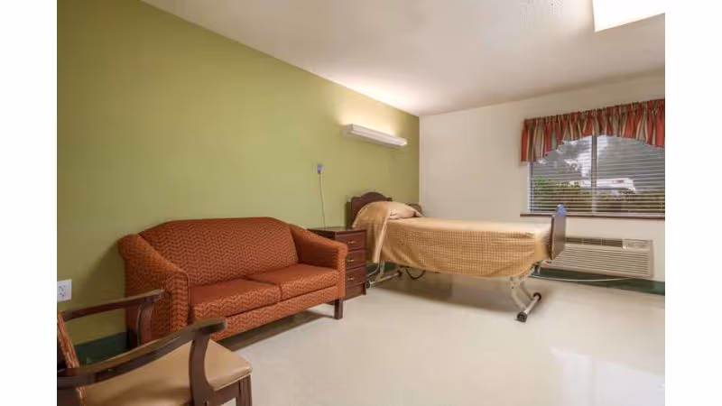 A simple room with a single hospital-style bed covered with a beige blanket, a red patterned couch, a wooden chair with a beige cushion, a small wooden nightstand, and a window with blinds and a red valance. The walls are painted green and white, and there is a wall-mounted light fixture above the bed.