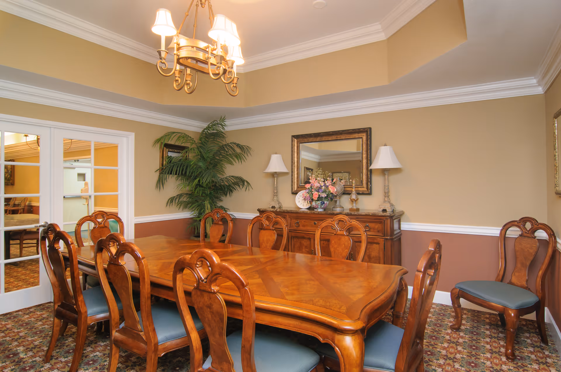 A formal dining room with a large wooden dining table surrounded by eight matching wooden chairs with blue cushions. The room has beige and brown walls with white trim, a chandelier hanging from the ceiling, a large mirror above a wooden sideboard decorated with lamps and a floral arrangement, and a potted plant in the corner. French doors lead to another room.
