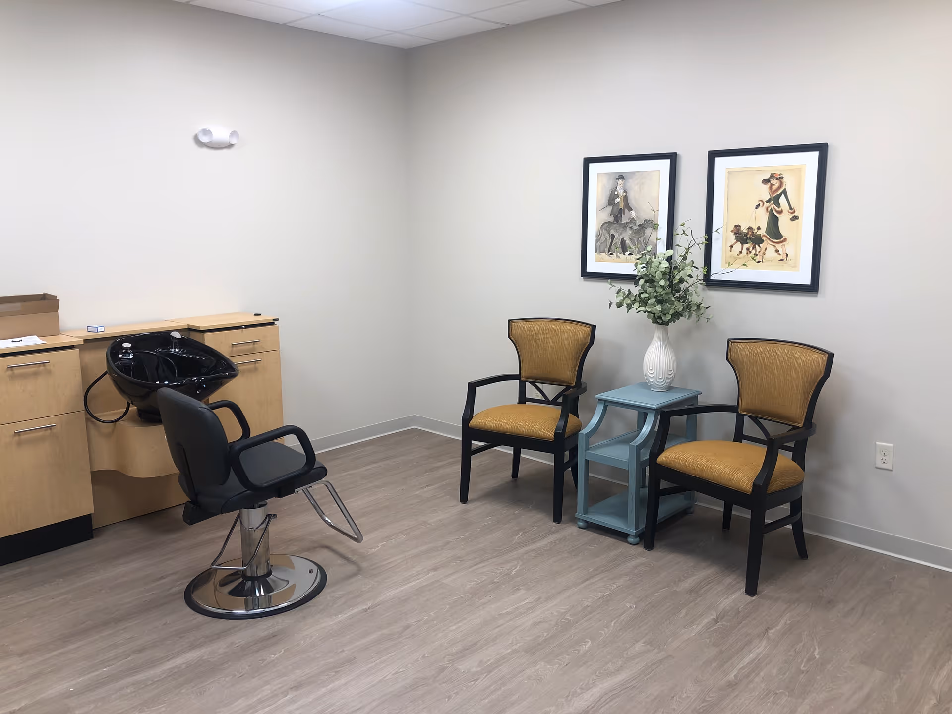 A small salon area with a black salon chair in front of a black hair washing sink attached to a wooden cabinet. Two yellow cushioned chairs with black frames are placed next to a small blue side table holding a white vase with green foliage. Two framed pictures hang on the wall above the chairs. The room has light-colored walls and wood flooring.