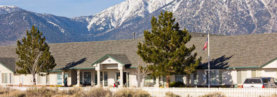 Single-story building with a gray shingled roof and white exterior walls, surrounded by trees and a white picket fence, set against a backdrop of snow-capped mountains under a clear blue sky.
