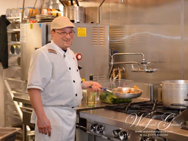 A chef wearing a white uniform and beige cap is cooking in a commercial kitchen, holding a pan with vegetables over a stove. The kitchen has stainless steel appliances and shelves with plates in the background.
