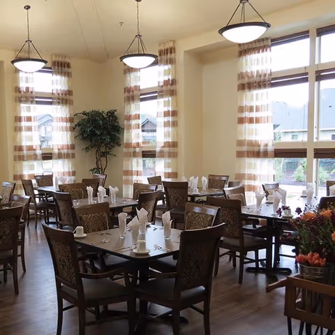 Bright dining room with multiple tables and chairs set with napkins, large windows with striped curtains, pendant lights, and a potted plant.