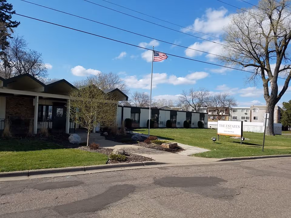 Front entrance of The Estates at Fridley senior living facility with a low-rise building, American flag on a flagpole, lawn and a sign.