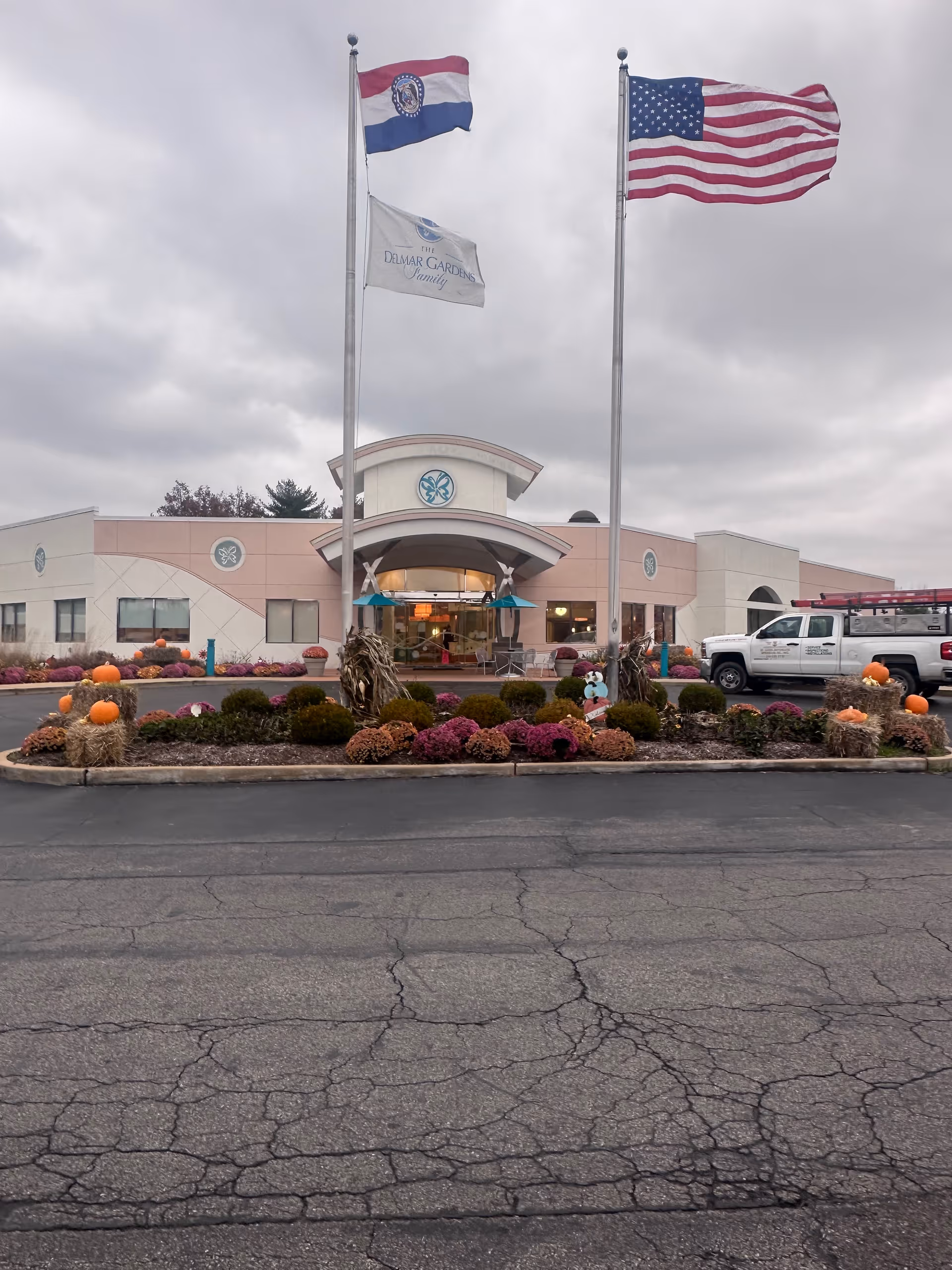 Exterior view of a senior living facility with two flagpoles displaying the Missouri state flag, a white flag with 'The Delmar Gardens Family' logo, and the American flag. The building has a curved entrance with a covered awning and decorative landscaping featuring pumpkins and flowers in front. A white truck is parked to the right of the entrance under a cloudy sky.