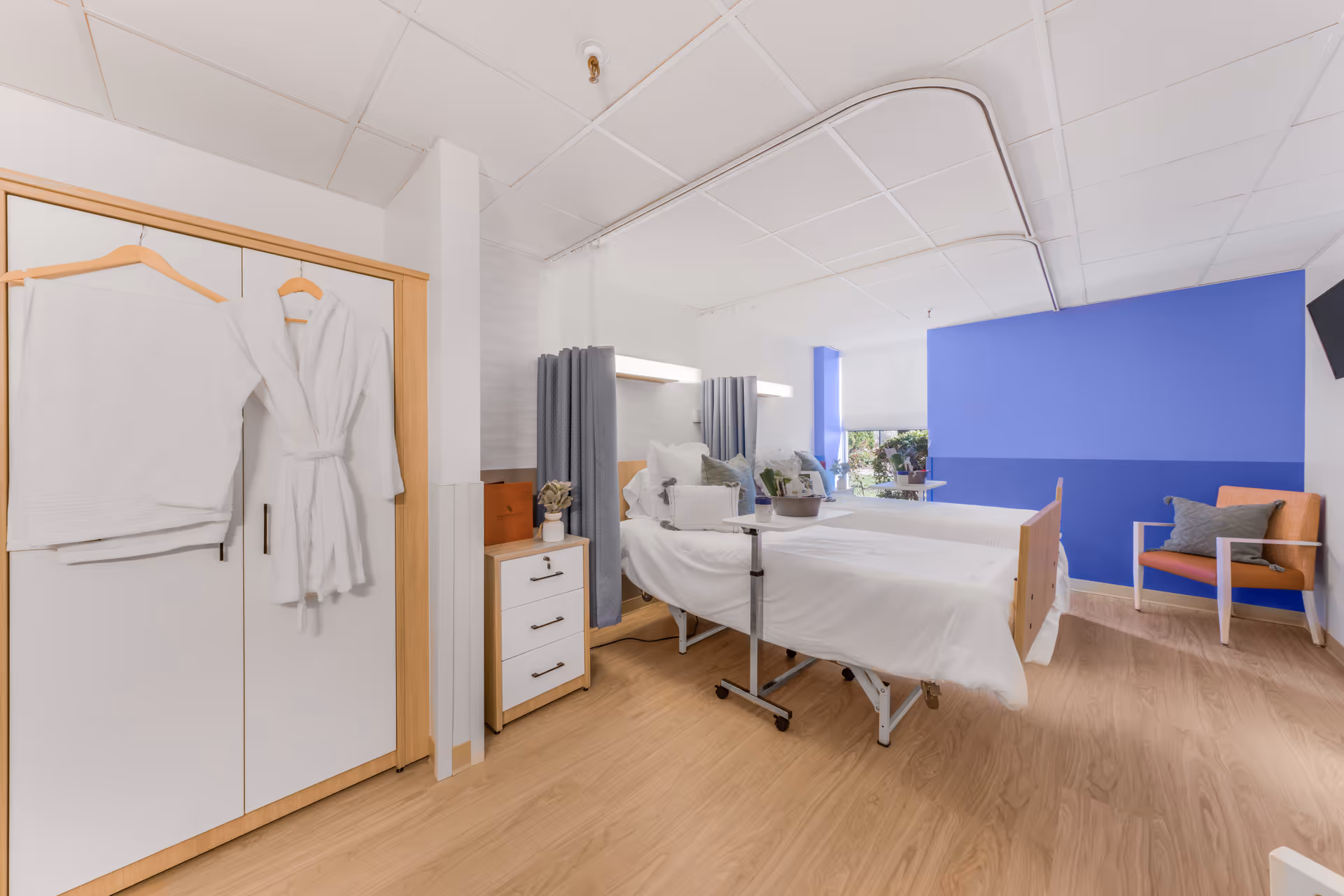 A bright and clean rehabilitation room with two hospital beds separated by a curtain. Each bed is made with white linens and has pillows. There is a small bedside cabinet with a plant and a brown box on it. On the left side, a wardrobe with a white towel and a white bathrobe hanging on the doors. The room has light wooden flooring, a blue accent wall, a window with a view of greenery, and an orange chair with a gray cushion near the wall.