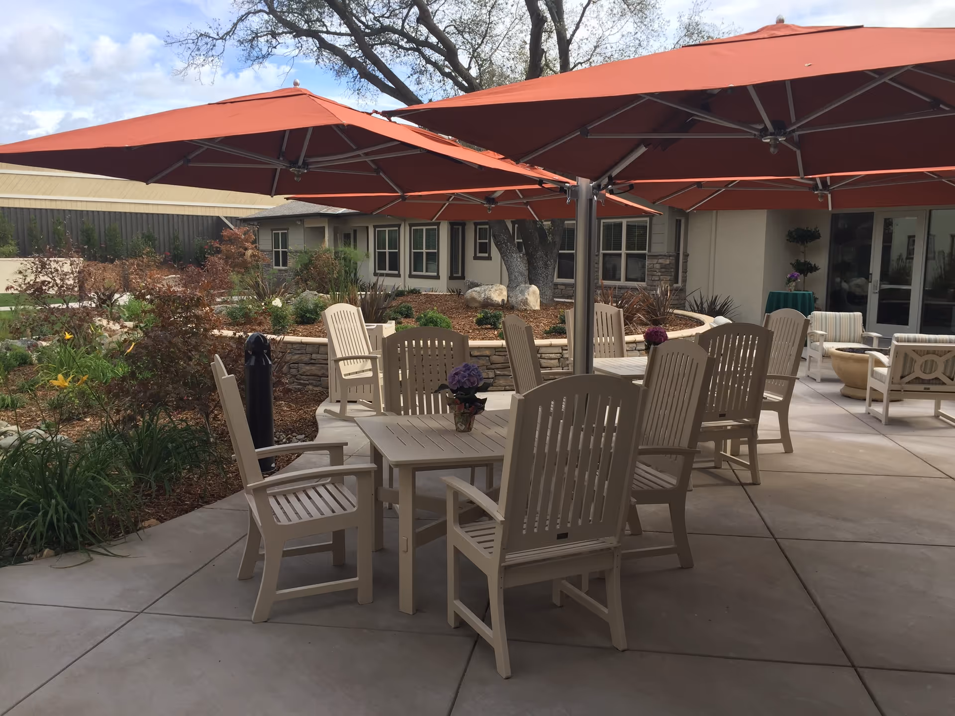 Outdoor patio area with beige tables and chairs under large red umbrellas, surrounded by landscaped garden beds and a building in the background.