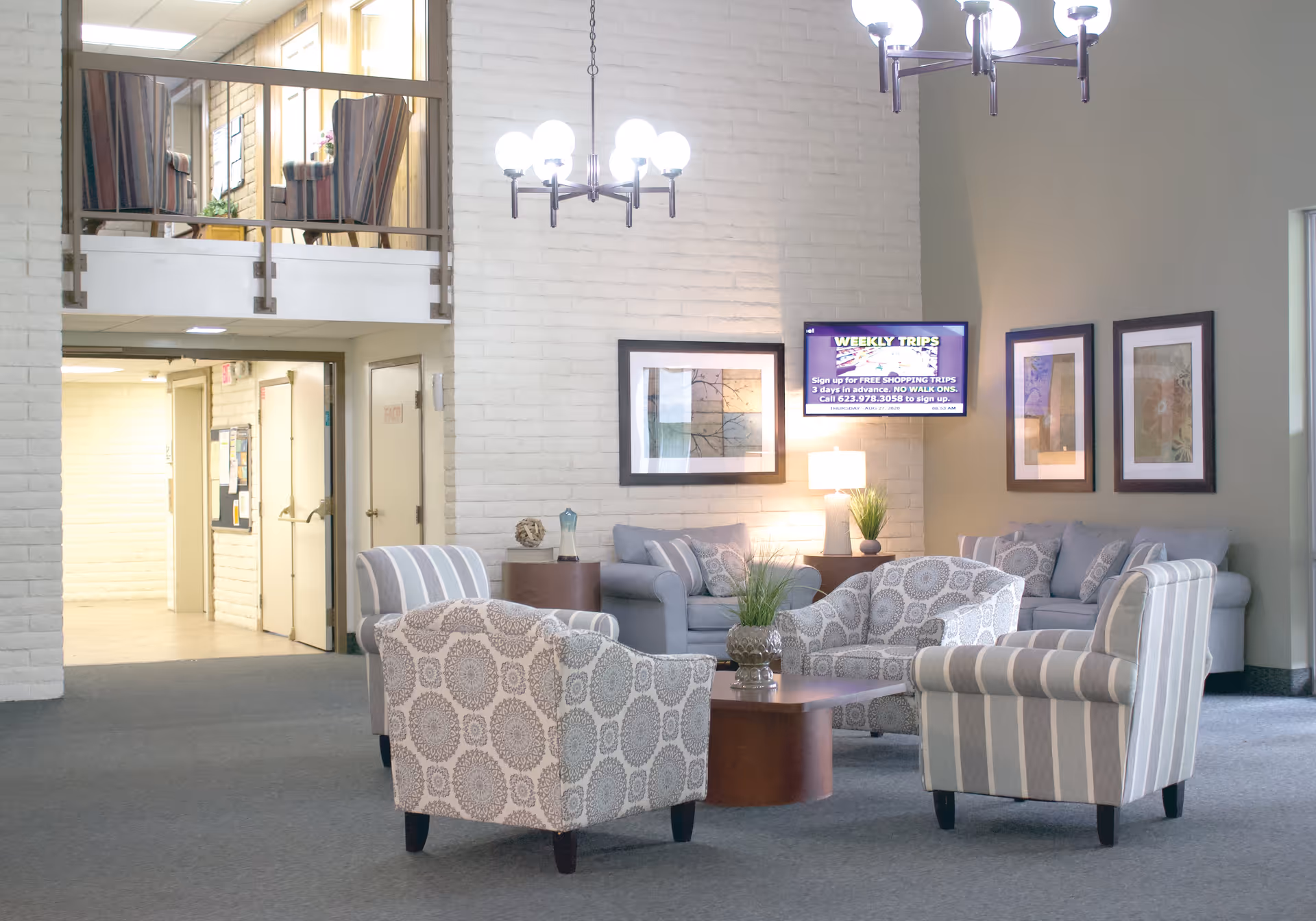 A bright and spacious living room area in a senior living facility featuring multiple patterned armchairs and sofas arranged around a wooden coffee table. The walls are light-colored with framed artwork and a mounted TV displaying information about weekly trips. There are two modern chandeliers hanging from the ceiling and a small plant on the coffee table. A hallway and an upper balcony with additional seating are visible in the background.