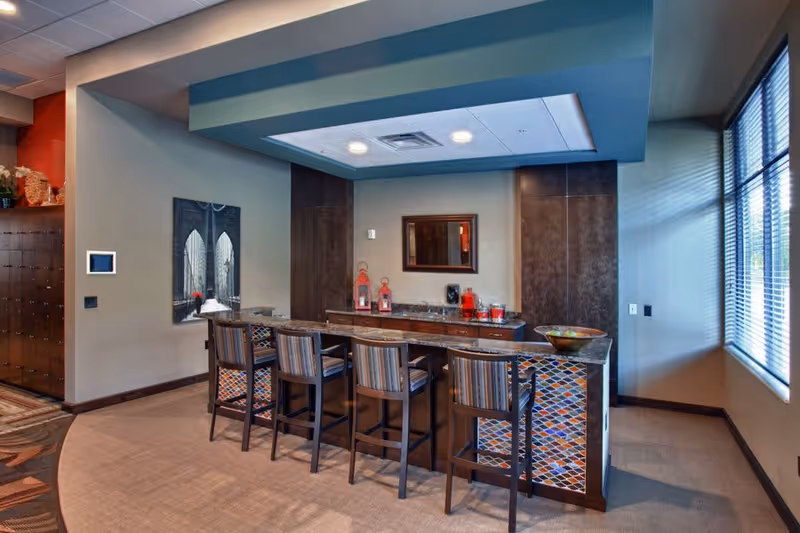 A bright senior living common area with a decorative bar counter, four high stools, and large windows.