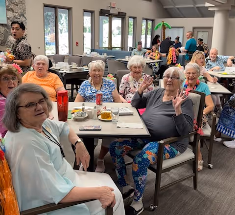 A group of elderly women sitting around a table in a bright dining area, smiling and waving at the camera. The room has large windows and several other people seated at tables in the background.