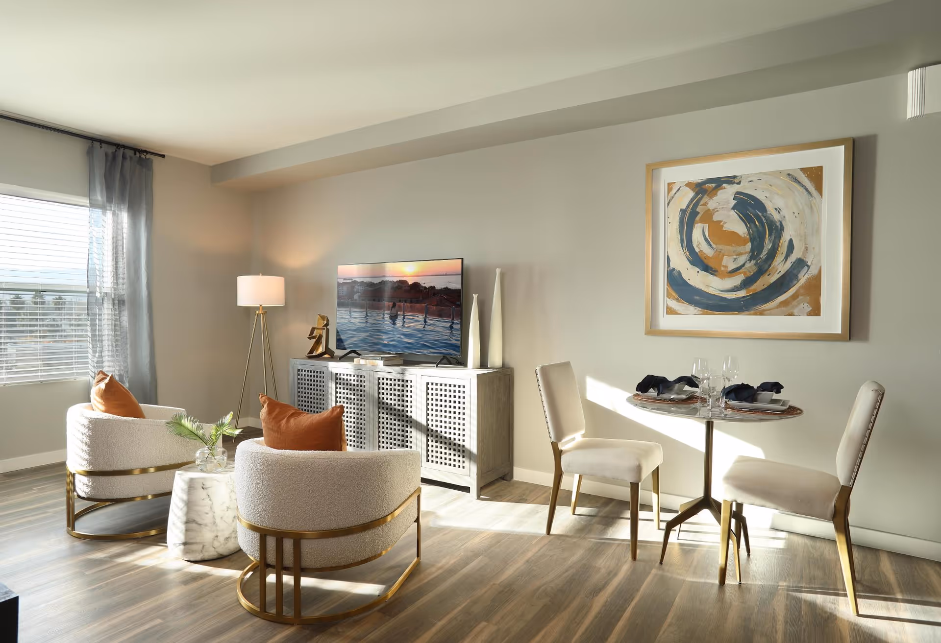 Bright modern living and dining area with two curved armchairs, a TV console, a small round dining table with two chairs, and abstract wall art.