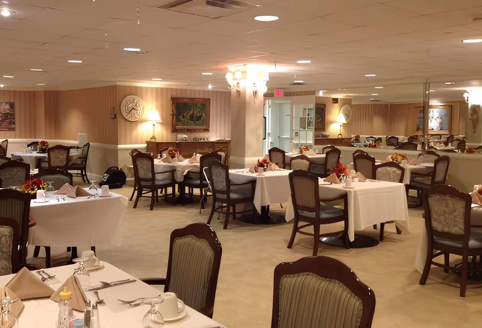 A well-lit dining room in a senior living facility with multiple tables covered in white tablecloths, set with napkins, glasses, and cutlery. The room features cushioned chairs, wall lamps, framed paintings, and a large mirror along one wall, creating a warm and inviting atmosphere.