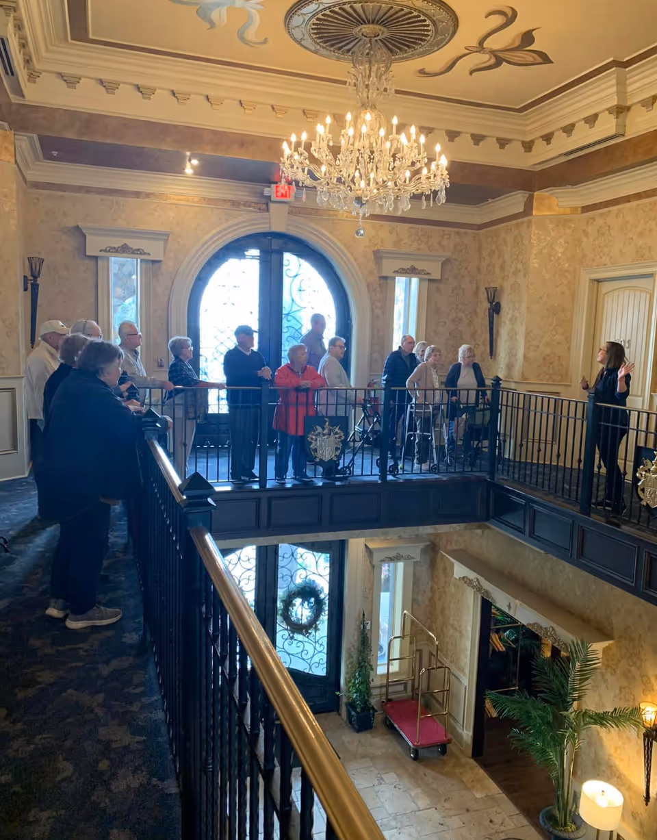 A group of elderly people standing on an upper balcony inside an elegant senior living facility, listening to a woman who appears to be giving a tour or presentation. The interior features ornate wallpaper, a large chandelier, decorative ceiling details, and a wrought iron railing overlooking a lower level with a luggage cart and potted plants.