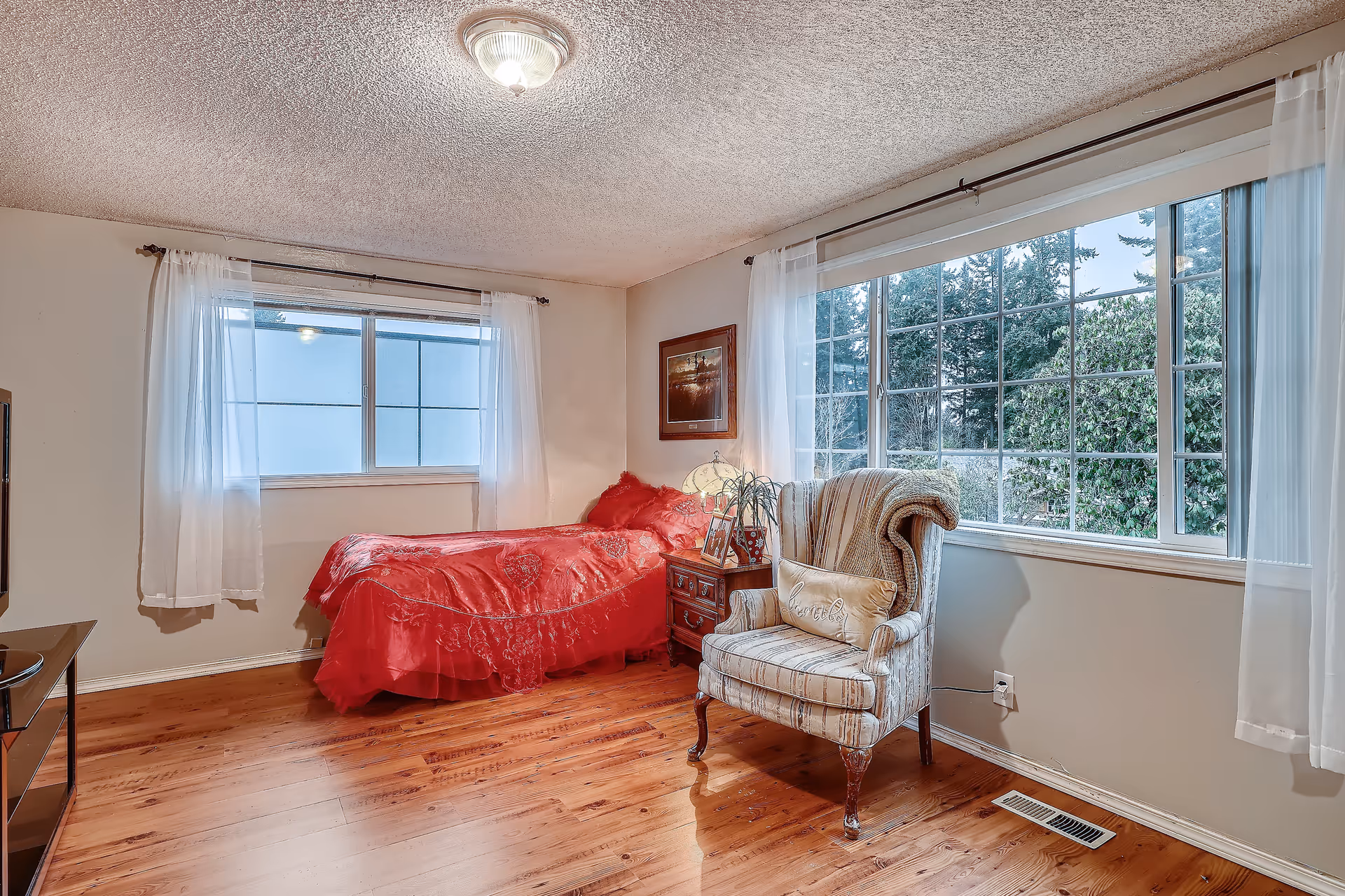 Sunny bedroom with a single bed covered in red bedding, an upholstered armchair, large windows and hardwood floors.