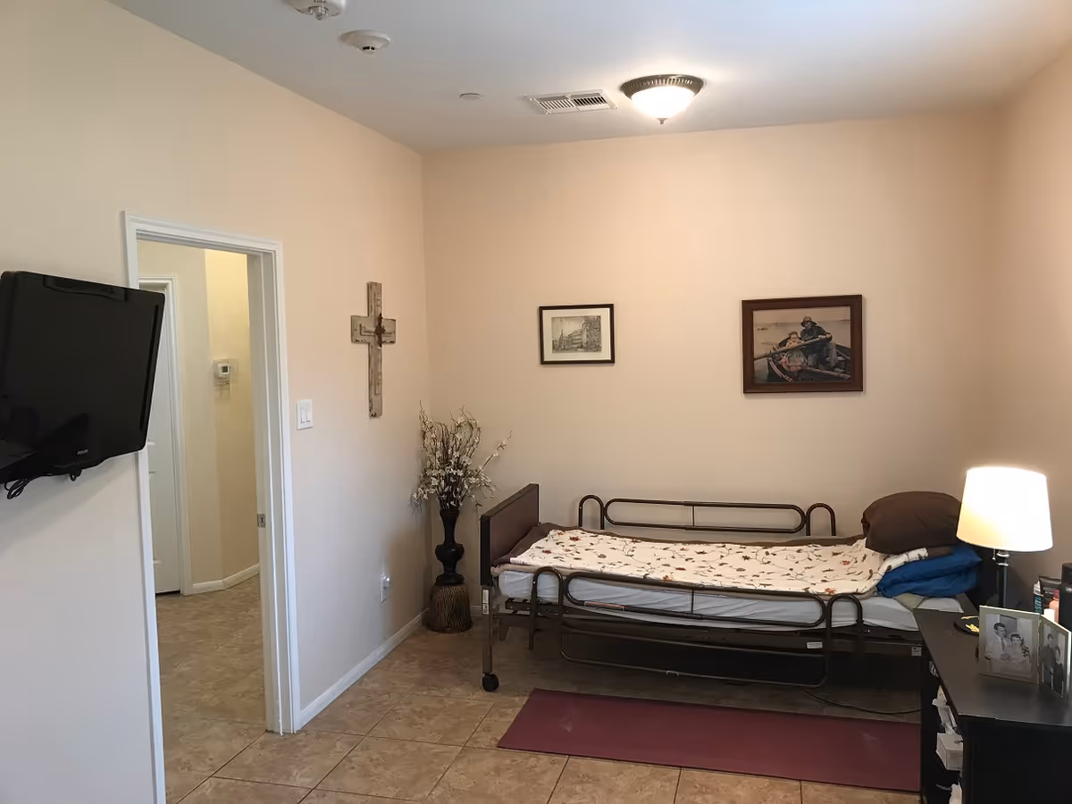A small bedroom with a single bed that has a metal frame and a floral bedspread. There is a bedside table with a lamp and framed photos on it. On the wall above the bed are two framed pictures. A decorative cross hangs on the adjacent wall near the doorway. A flat-screen TV is mounted on the wall to the left. The floor is tiled and there is a vase with dried flowers in the corner.