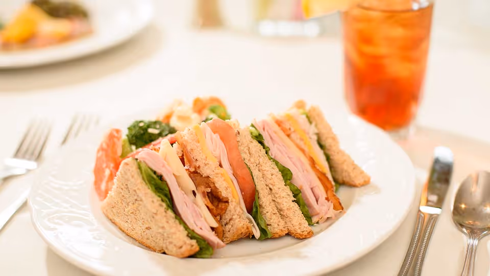 A close-up of a plate with club sandwiches made with whole wheat bread, lettuce, tomato, turkey, cheese, and bacon, served with a side of salad and a glass of iced tea in the background.