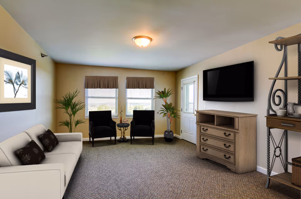 Furnished common living room with a sofa, two armchairs by windows, a wall-mounted TV above a dresser, and decorative shelving.