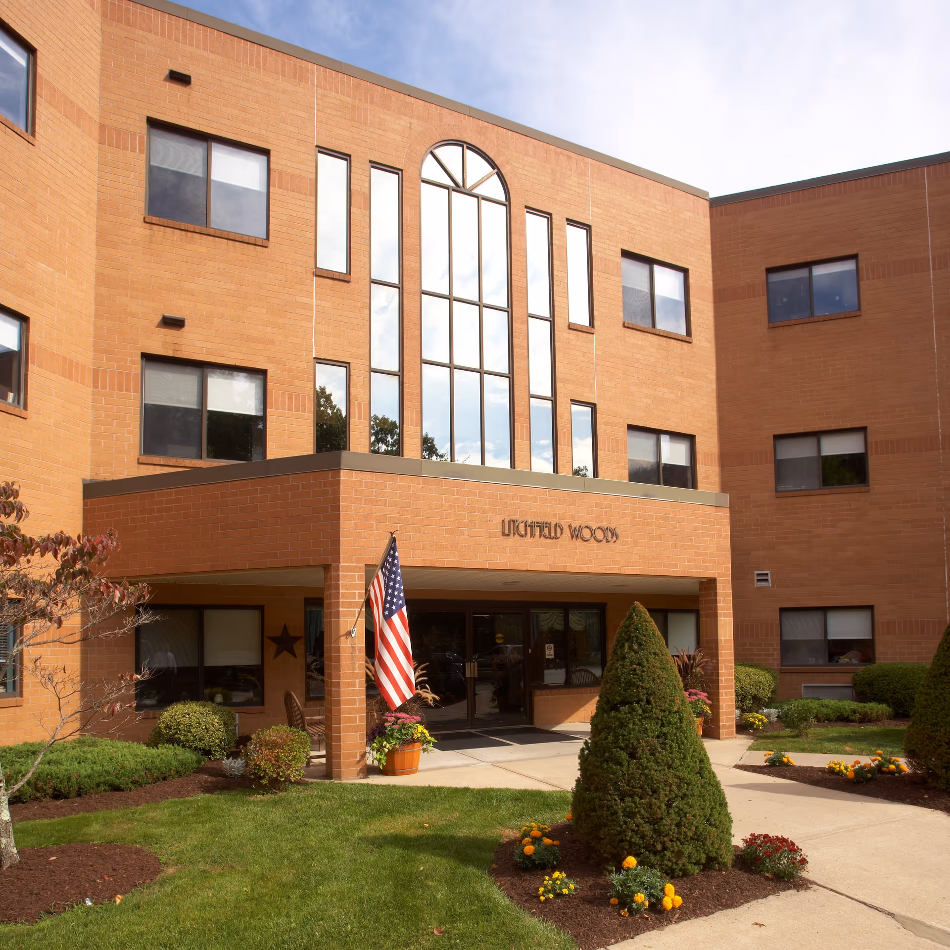 Exterior front view of a three-story brick building with large vertical windows above the entrance. The entrance has a covered porch with an American flag and potted plants. There are neatly trimmed bushes, a small tree, and flower beds in front of the building. The building has the name 'Litchfield Woods' displayed above the entrance.