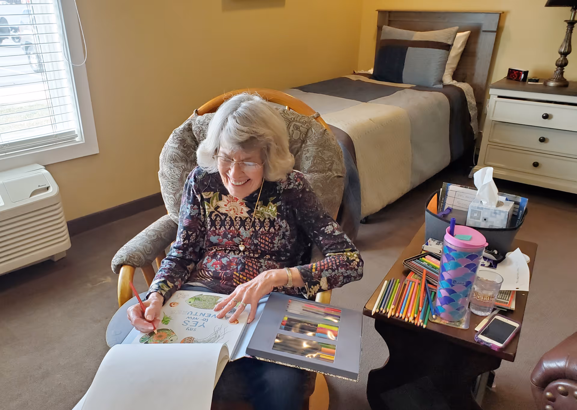 An elderly woman with gray hair and glasses is sitting in a cushioned rocking chair in a bedroom. She is smiling and coloring in a book with colored pencils. Next to her is a small table holding various colored pencils, a purple and blue patterned tumbler, a glass of water, a smartphone, and a container with tissues and other items. Behind her is a neatly made bed with a beige and gray comforter, a nightstand with a lamp, and a window with blinds letting in natural light.