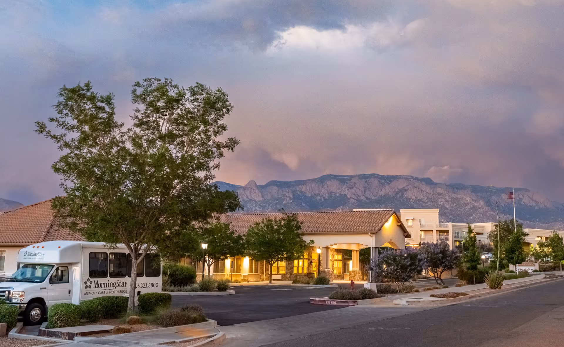 Front exterior of the MorningStar at North Ridge senior living facility with a shuttle van parked outside and mountains in the background at dusk.