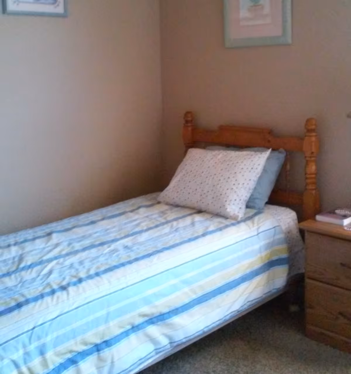 A small bedroom with a single wooden bed featuring a striped blue, yellow, and white bedspread and two pillows. Next to the bed is a wooden nightstand with a few books and a small item on top. The walls are beige, and there are two framed pictures hanging above the bed.
