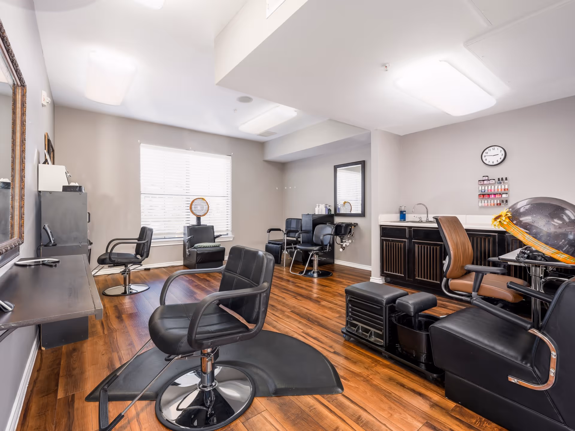 Interior view of a salon room with multiple black salon chairs, a hair dryer chair, a large mirror, a sink with cabinets underneath, and a window with blinds letting in natural light. The room has wooden flooring and neutral-colored walls.