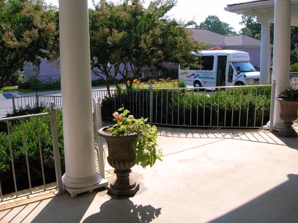 Covered front porch with columns and planters overlooking a landscaped driveway and a shuttle bus marked The Yellow Brick House.