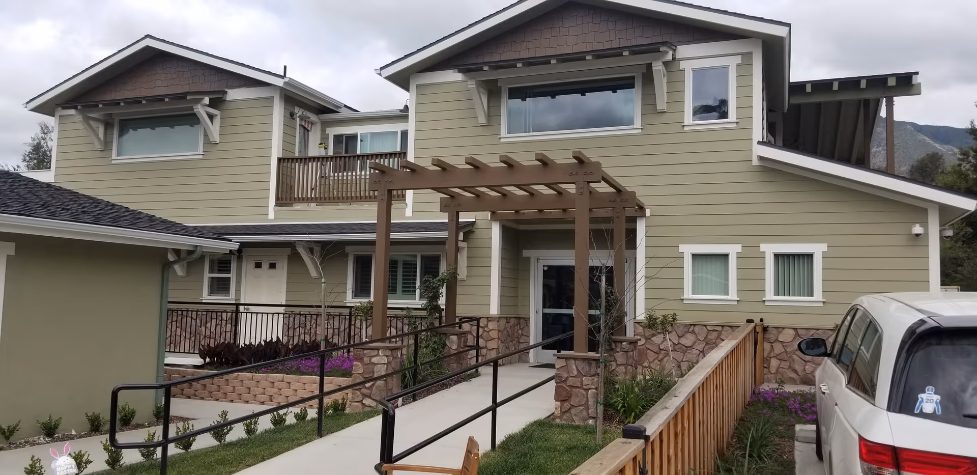 Exterior view of a two-story senior living facility building with green siding and white trim. There is a stone base around the lower part of the building, a wooden pergola over the entrance walkway, and a ramp with black handrails leading to the door. A white vehicle is parked on the right side, and there are plants and flowers along the walkway and building.