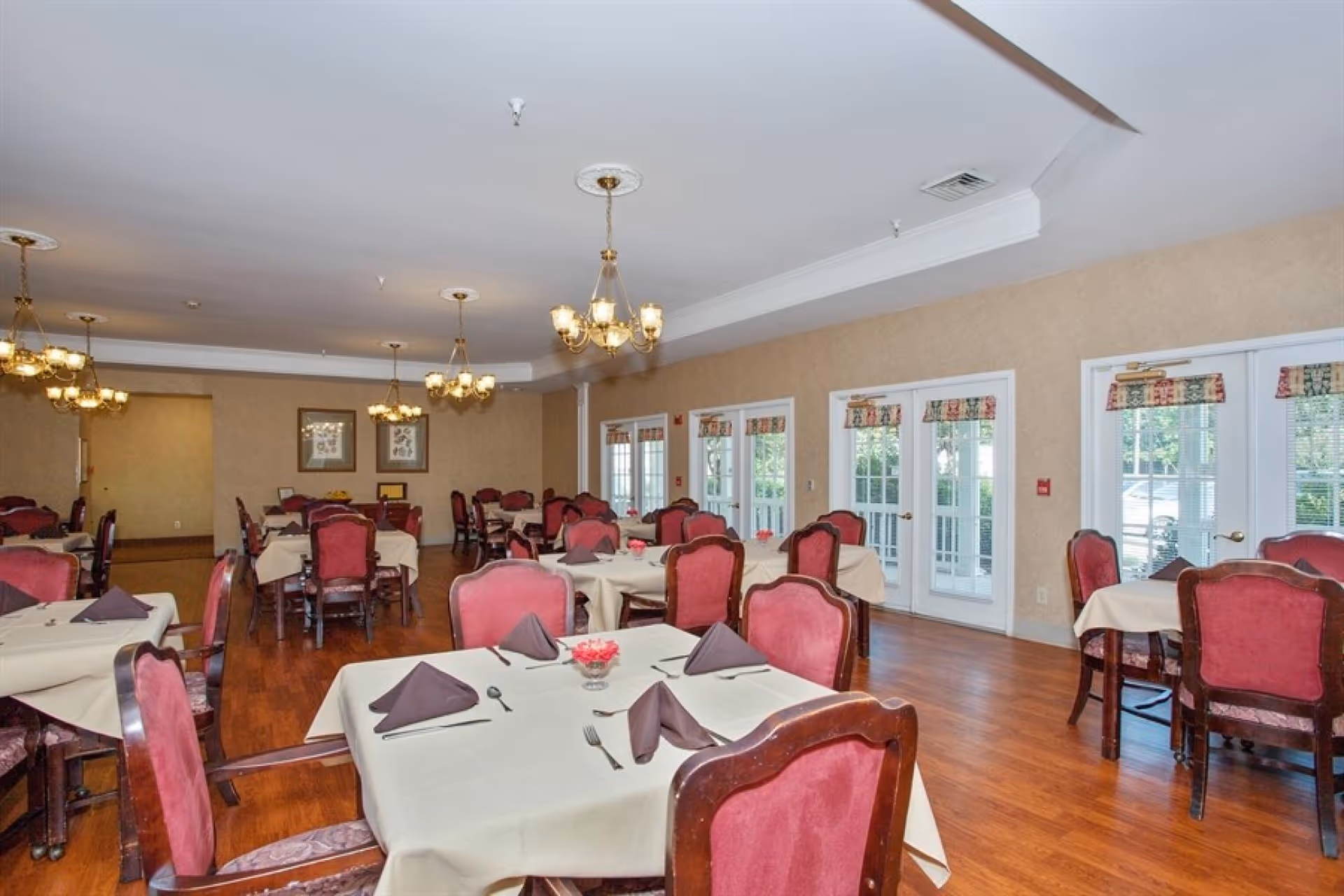 A dining room with multiple tables covered in beige tablecloths, each set with brown folded napkins, silverware, and small flower arrangements. The chairs have red upholstery and wooden frames. The room has wooden flooring, beige walls, several chandeliers hanging from the ceiling, and large windows with glass doors letting in natural light.