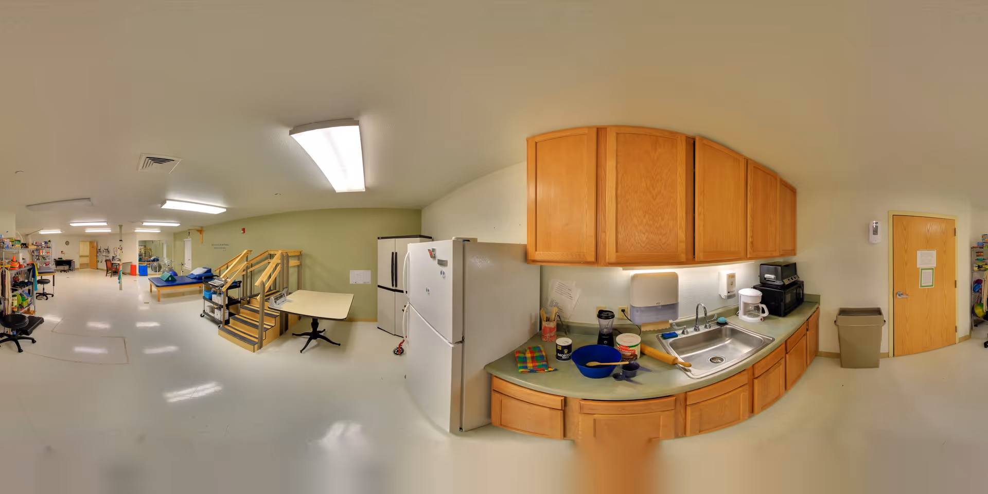A wide view of an interior room in a senior living facility showing a kitchenette area with wooden cabinets, a sink, a refrigerator, and various kitchen items on the counter. The room also contains exercise equipment, tables, chairs, and a set of stairs leading to a raised platform. The walls are light-colored, and the floor is white with overhead fluorescent lighting.
