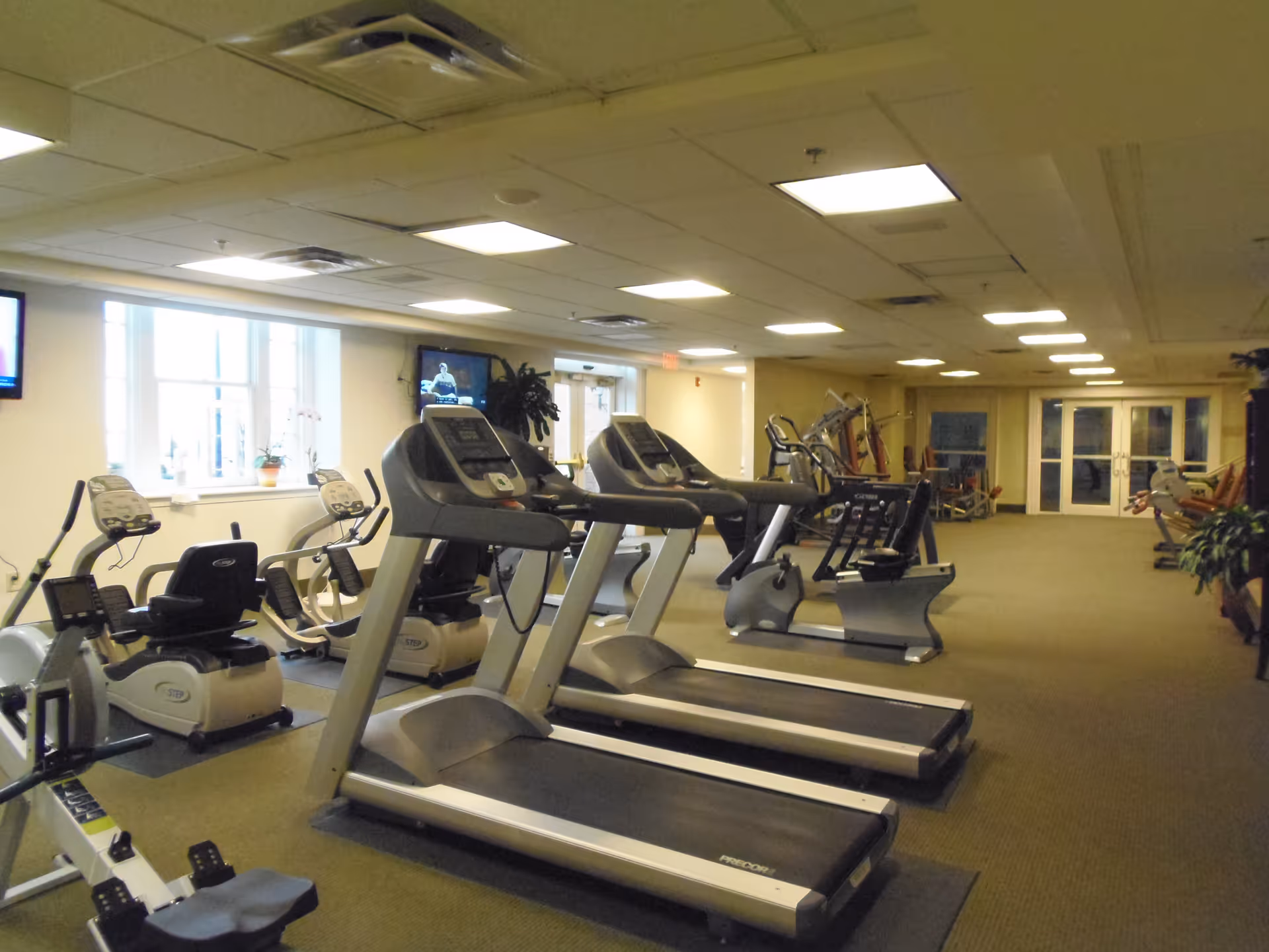 Interior view of a fitness room in a senior living facility with treadmills, exercise bikes, and other workout equipment arranged in a spacious, well-lit area with beige walls and carpeted floor.