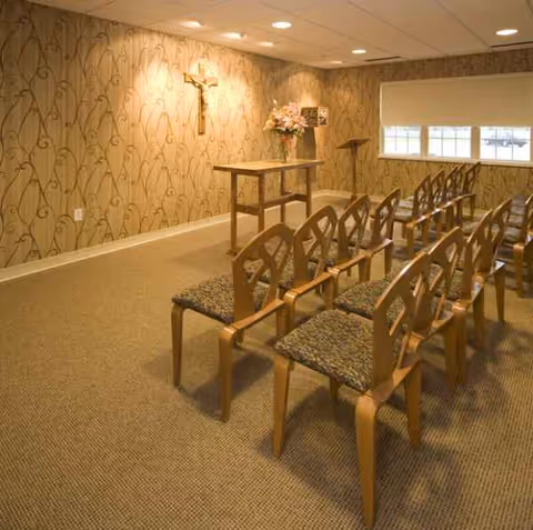 Chapel-like interior with rows of wooden chairs, a cross wall, and a small front table.