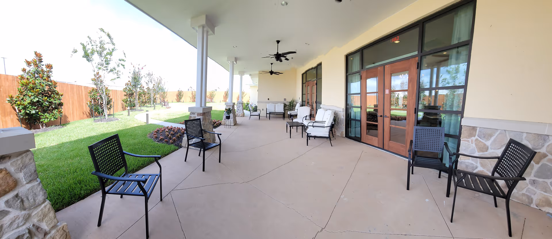 Covered outdoor patio area with multiple black metal chairs and cushioned seating. The patio has ceiling fans and stone pillars, overlooking a grassy garden area with small trees and a wooden fence.