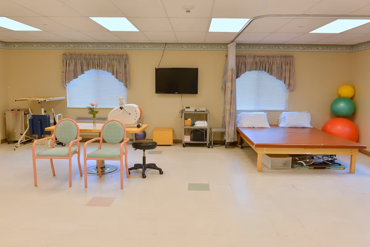 A therapy or rehabilitation room with two windows covered by patterned valances and blinds. There is a wooden table with two green cushioned chairs and a black rolling stool. On the table, there is a vase with flowers and a piece of exercise equipment. To the right, there is a large padded therapy table with two pillows and three large exercise balls stacked in the corner. A flat-screen TV is mounted on the wall above a small cart with towels and equipment.