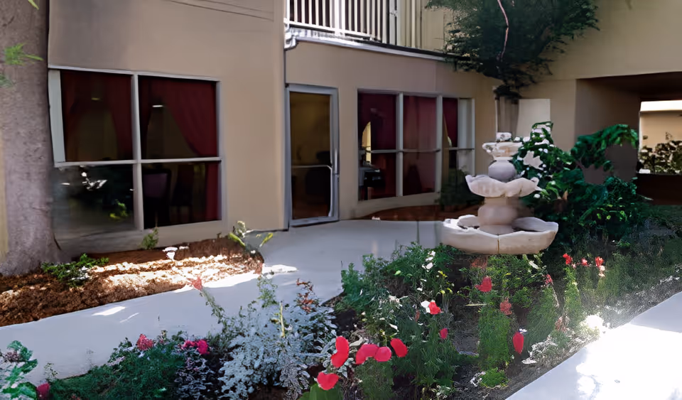 A small courtyard with a tiered stone fountain, flower beds, and windows and a door of the assisted living building.