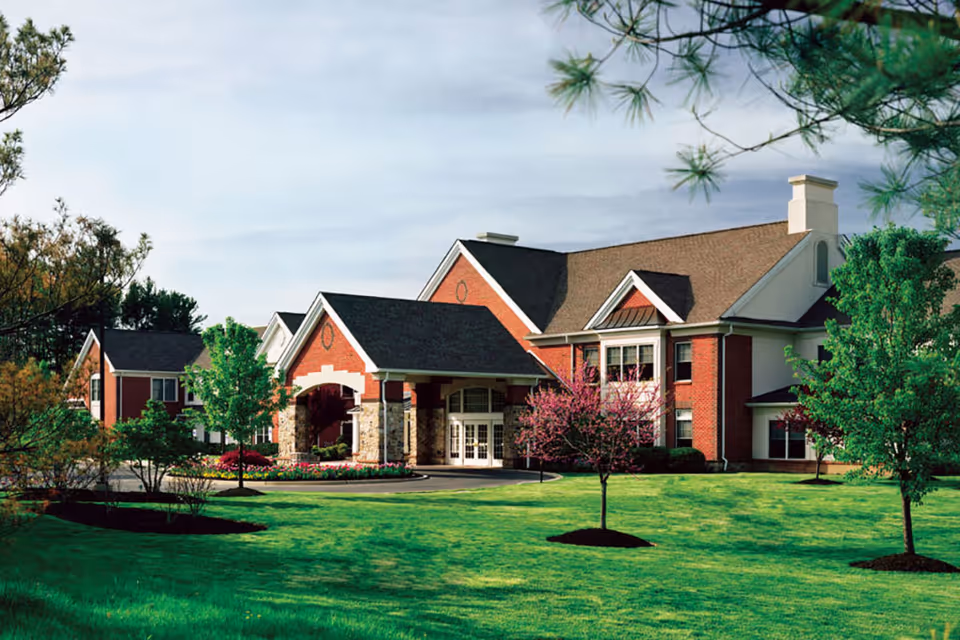 Exterior view of a large brick building with a covered entrance, surrounded by green grass, trees, and landscaping under a partly cloudy sky.