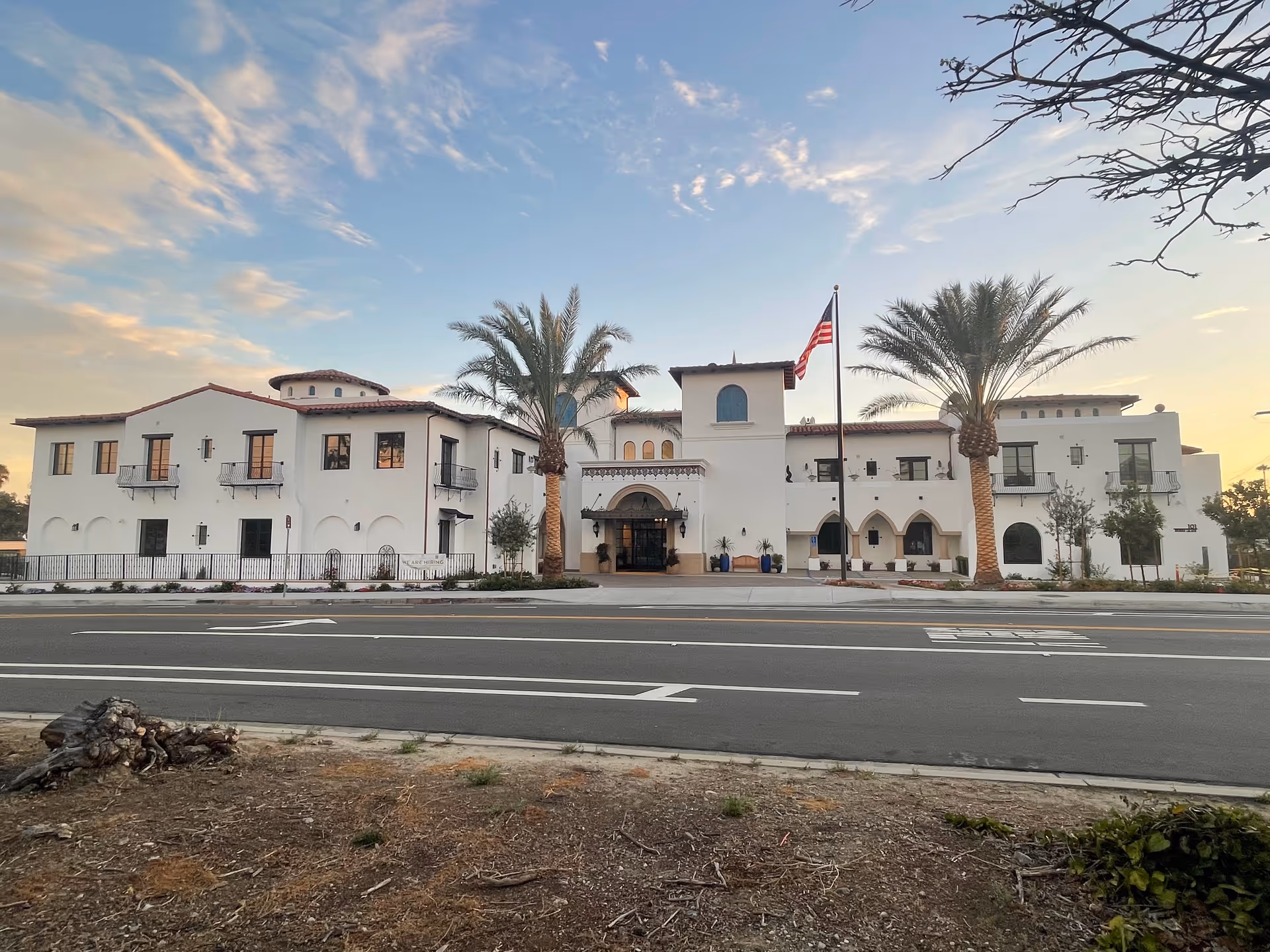 Front exterior view of a large white building with Spanish-style architecture, featuring arched windows and doorways, a tiled roof, two tall palm trees, and an American flag on a flagpole in front. The sky is partly cloudy with a warm light from the setting or rising sun.