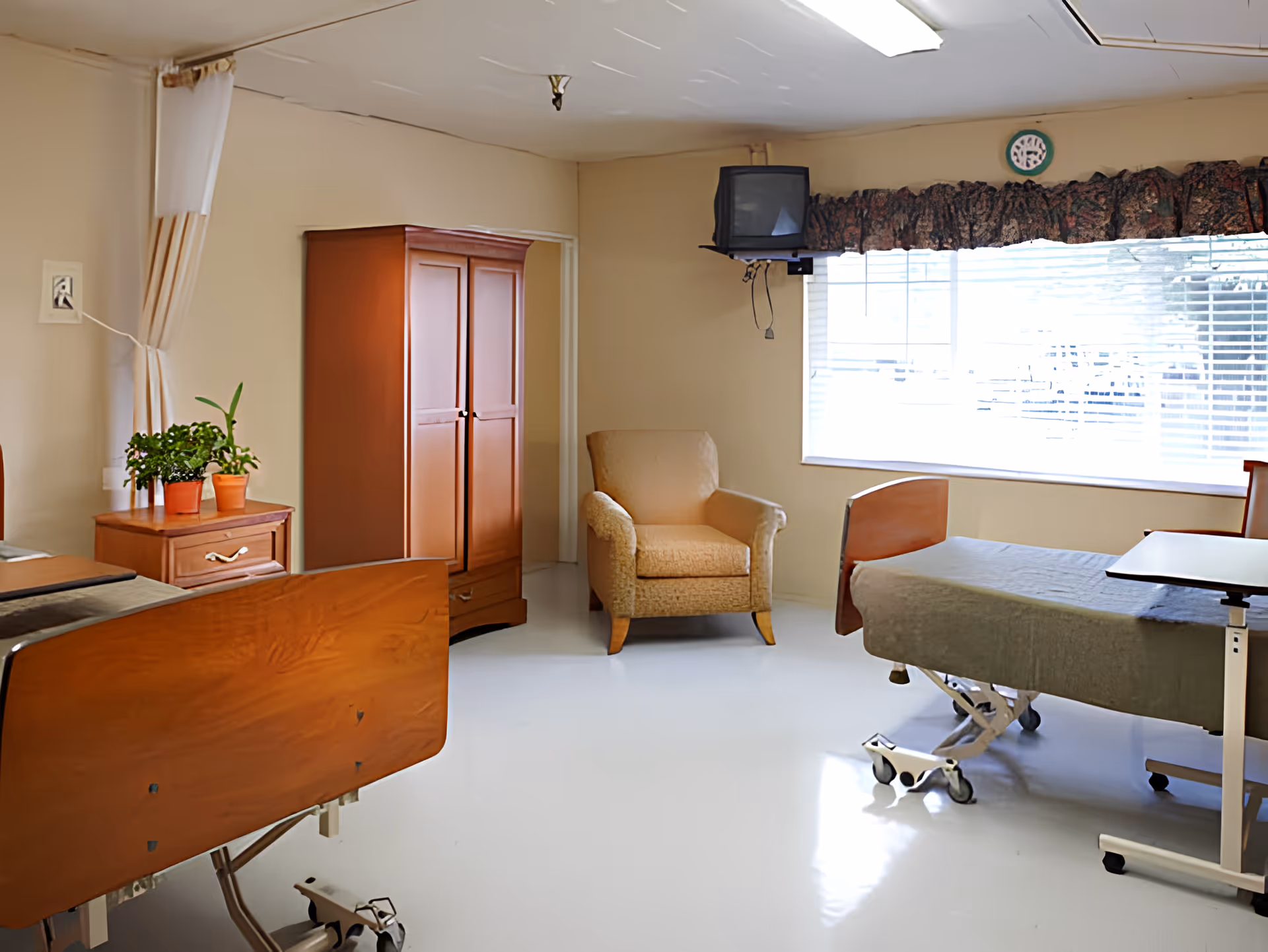 A rehabilitation room with two hospital beds, a wooden wardrobe, a small wooden nightstand with potted plants, a beige armchair, a wall-mounted TV, and a large window with blinds and a floral valance.