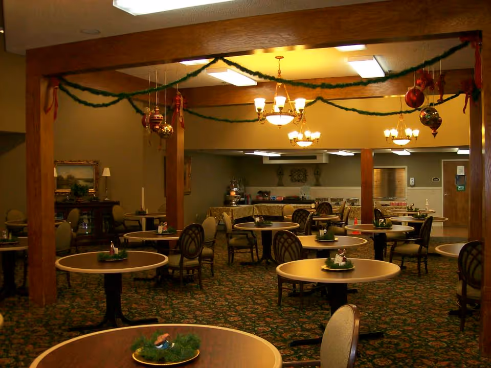 A dining room decorated for the holidays with green garlands and red ornaments hanging from wooden beams. The room contains multiple round tables with chairs arranged around them, each table having a small holiday centerpiece. The carpet has a floral pattern, and there are chandeliers providing warm lighting. In the background, there is a buffet area with covered dishes and a wooden sideboard with a lamp and framed artwork on the wall.
