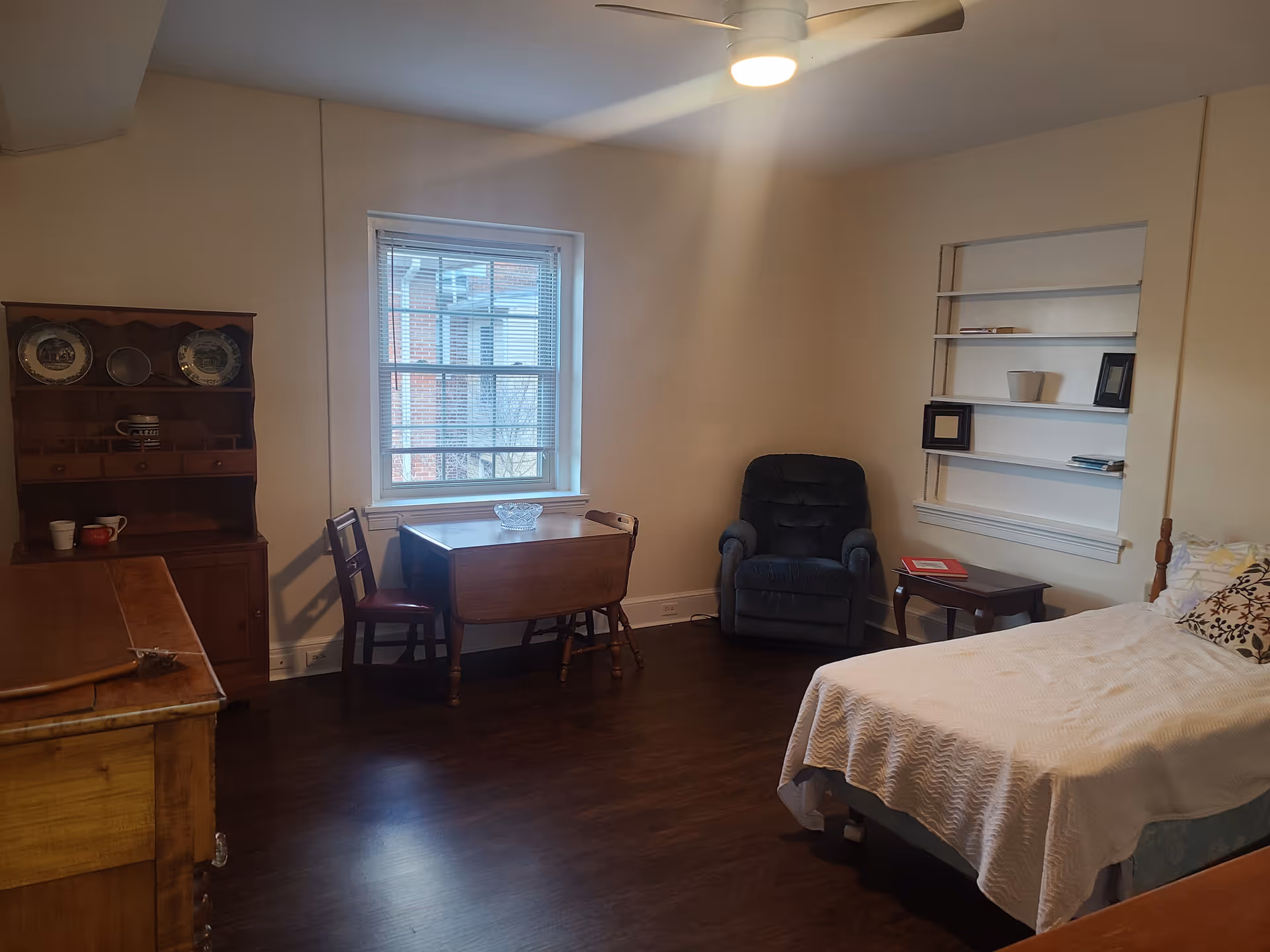 Well-lit bedroom with a bed, upholstered recliner, wooden dresser and a small table by a window.