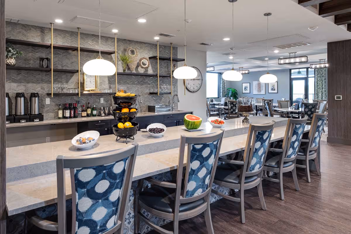 Communal dining and bar area with a long marble counter, patterned high-back chairs, pendant lights, and dining tables in the background.