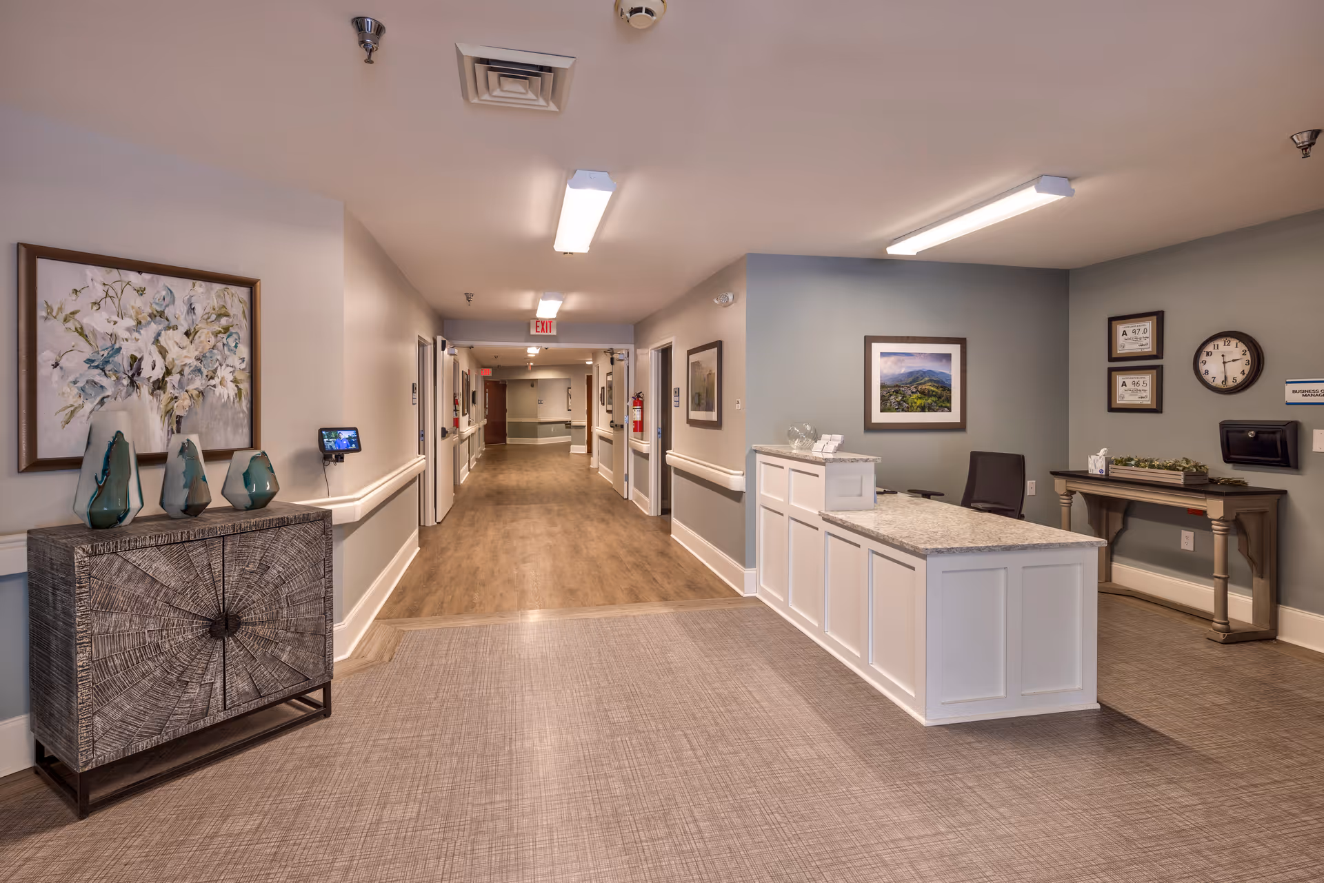 A clean and well-lit hallway in a senior living facility with a reception desk on the right side, decorated with framed pictures and a clock on the wall. On the left side, there is a decorative cabinet with three green vases and a large floral painting above it. The hallway extends into the distance with doors on both sides and wooden flooring.