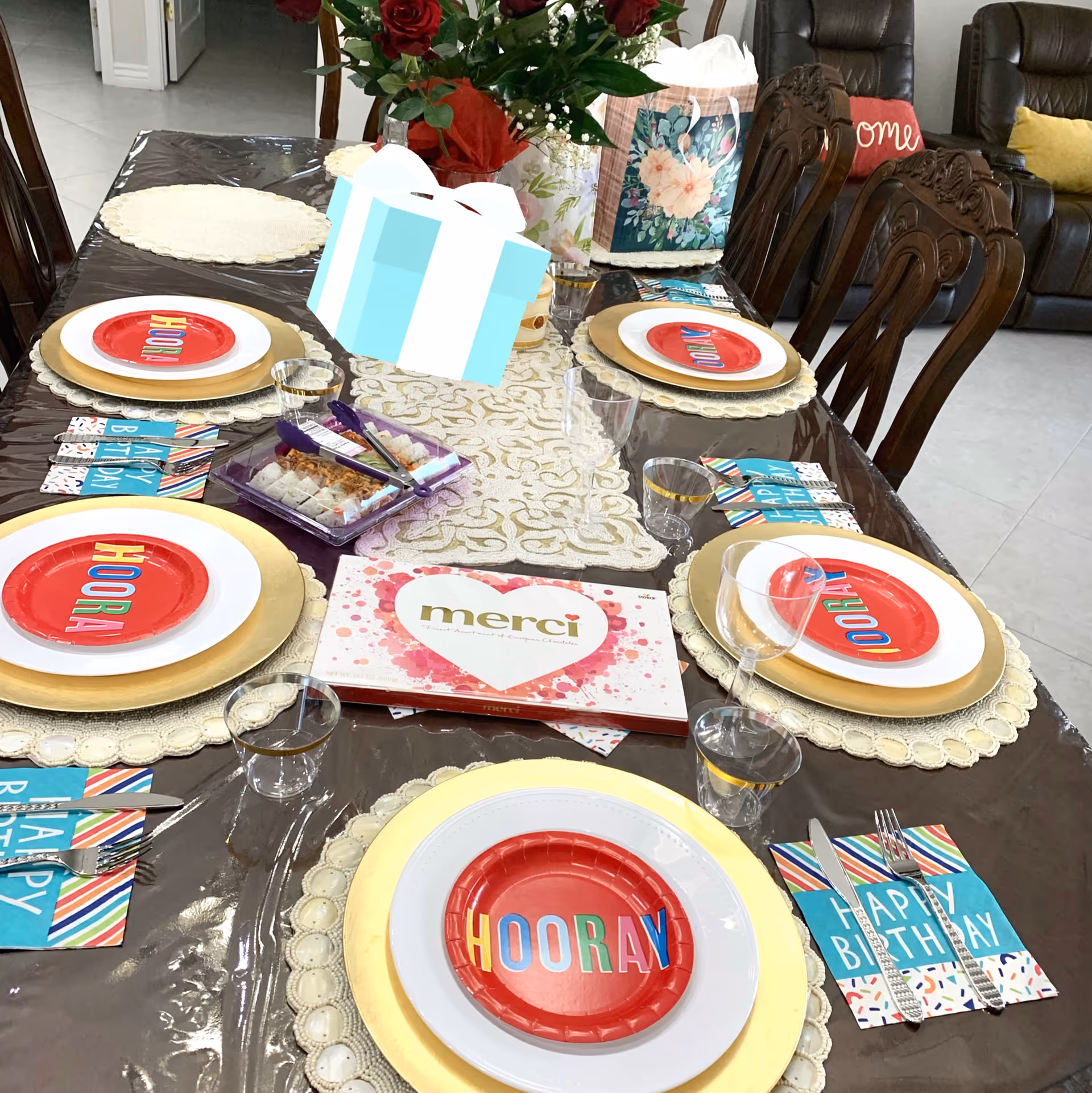 A dining table set for a celebration with plates, glasses, and colorful napkins that say 'HAPPY BIRTHDAY'. The plates have smaller red plates on top with the word 'HOORAY' in multicolored letters. There is a box of Merci chocolates and a floral gift bag on the table. In the background, there are brown leather chairs and a sofa with decorative pillows.