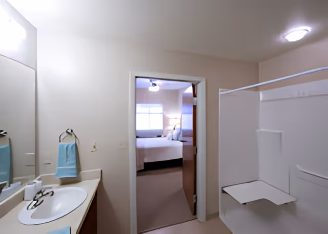 A bathroom with a white sink and countertop on the left, a mirror above the sink, and a blue hand towel hanging on the wall. On the right side, there is a white shower area with a fold-down seat. Through the open door, a bedroom with a bed and a window is visible.